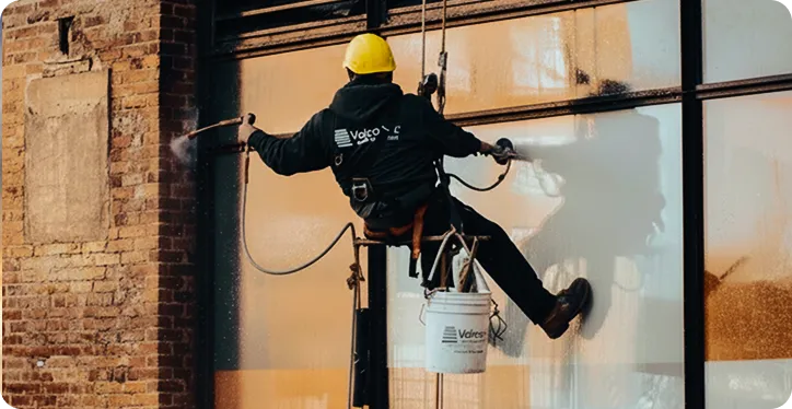 Rope-access worker pressure washing a glass facade on a brick building exterior.