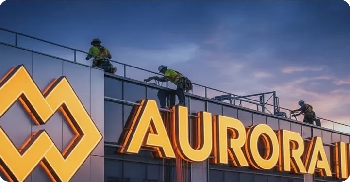 Rope-access workers performing maintenance on illuminated “Aurora” signage atop a commercial building at dusk.