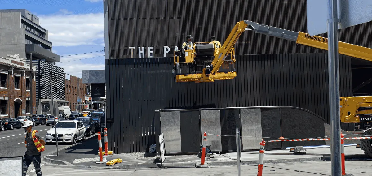 Workers using a boom lift to perform exterior maintenance on a commercial building along a city street.
