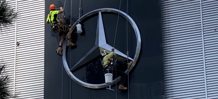 Rope-access workers cleaning a large Mercedes-Benz logo on a commercial building facade.
