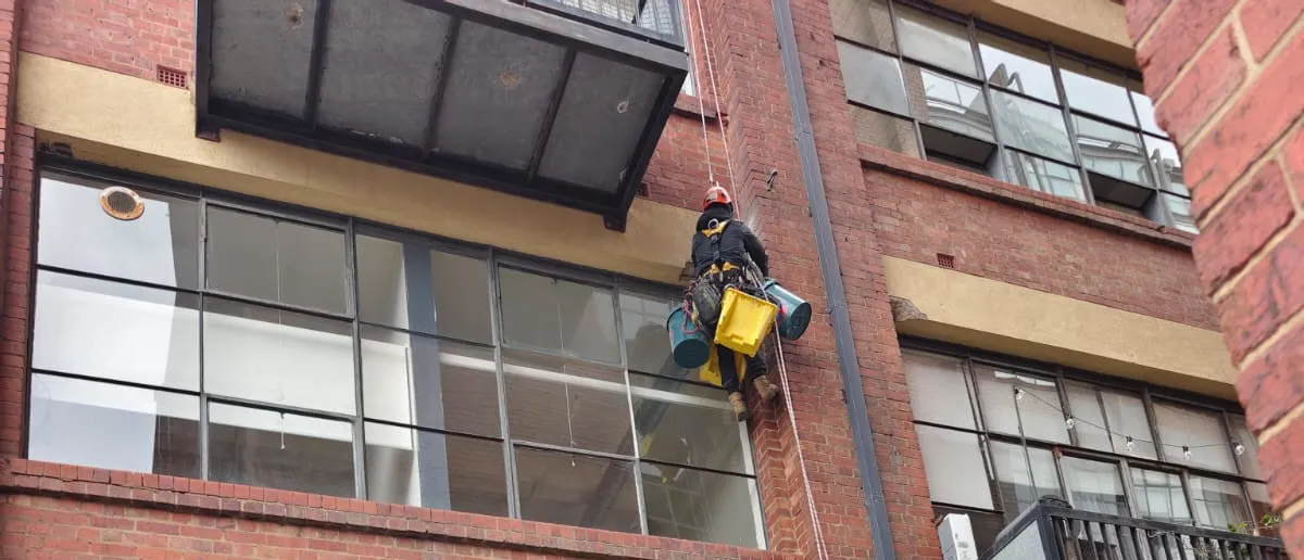 Service Rope-access technician cleaning windows on a brick building facade with suspended gear and buckets.